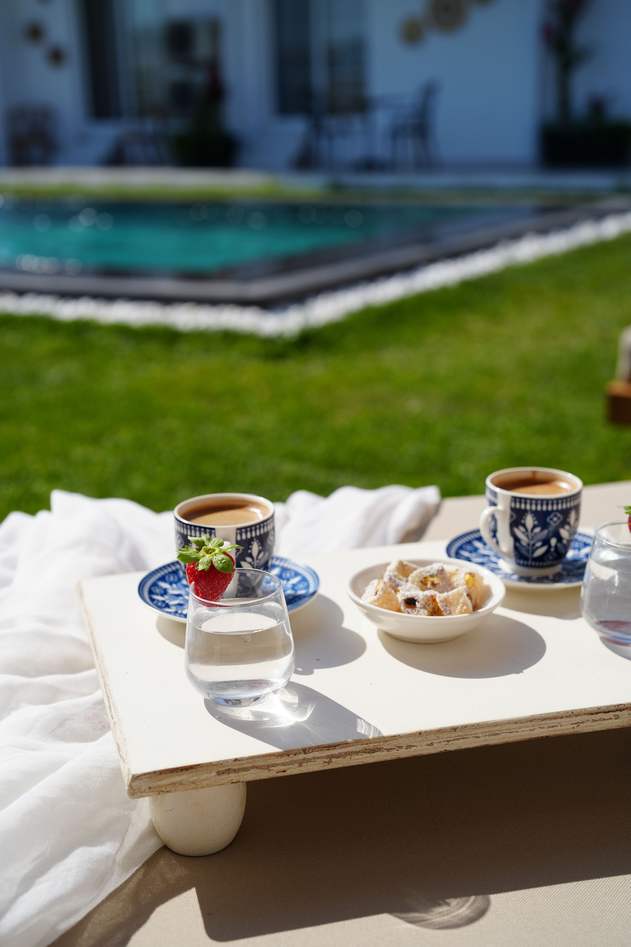 A table with a cup of coffee, a glass of water, a plate of pastries, and a cup of tea, set on a white cloth outdoors.