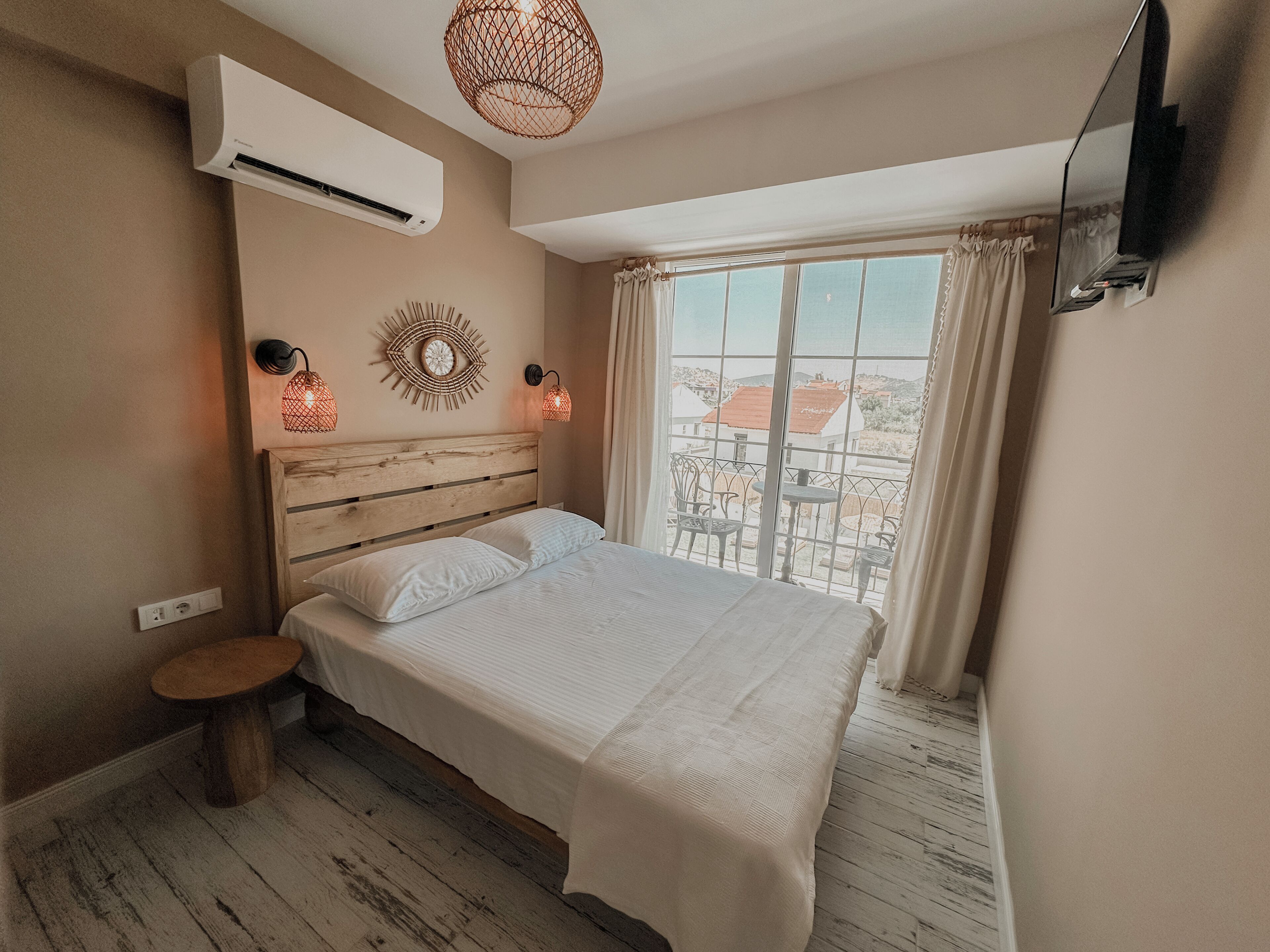 Un dormitorio con cama, aire acondicionado, una mesita, televisor montado en la pared y vistas a edificios a través de una ventana.