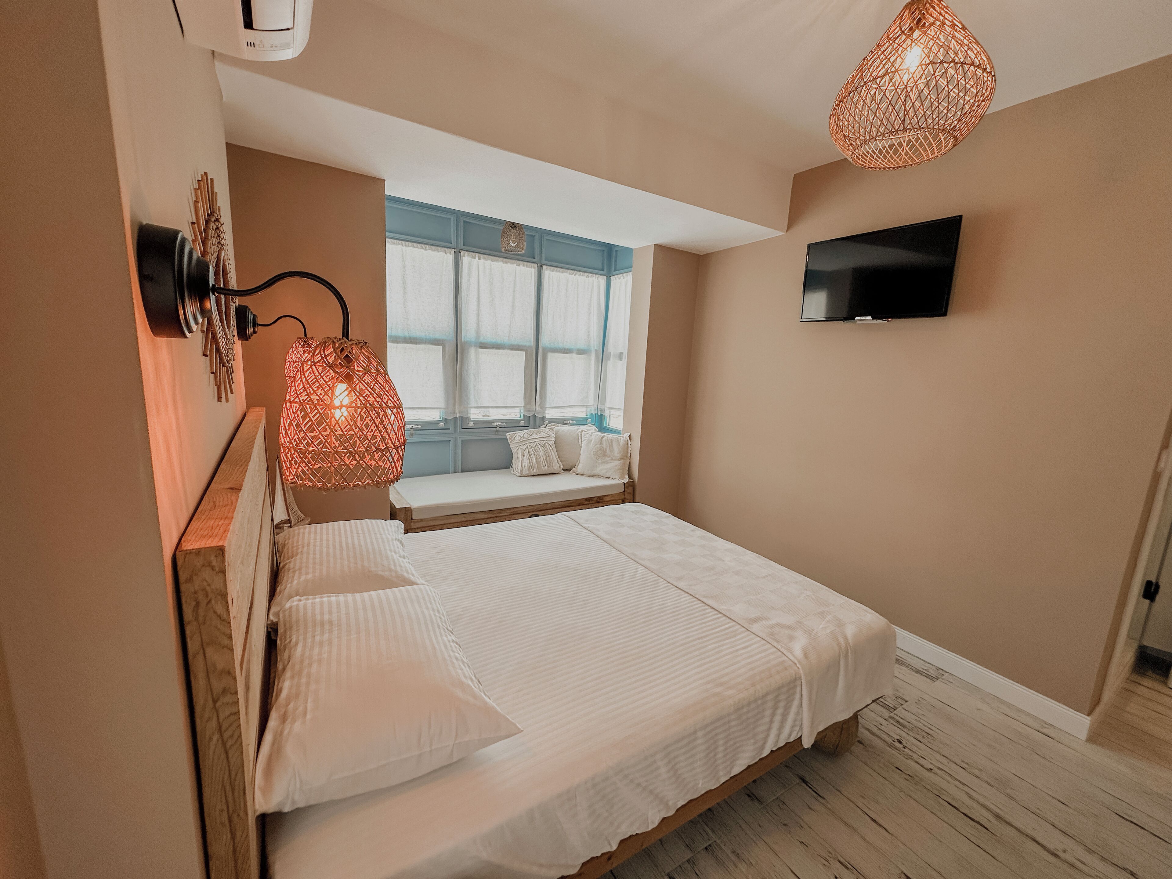 A bedroom with a bed, a TV mounted on the wall, a window with curtains, and a hanging light fixture.