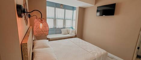 A bedroom with a bed, a TV mounted on the wall, a window with curtains, and a hanging light fixture.