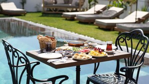 A poolside dining table with a variety of food and drinks, surrounded by lounge chairs and umbrellas. - Villa Egella (Dikili)