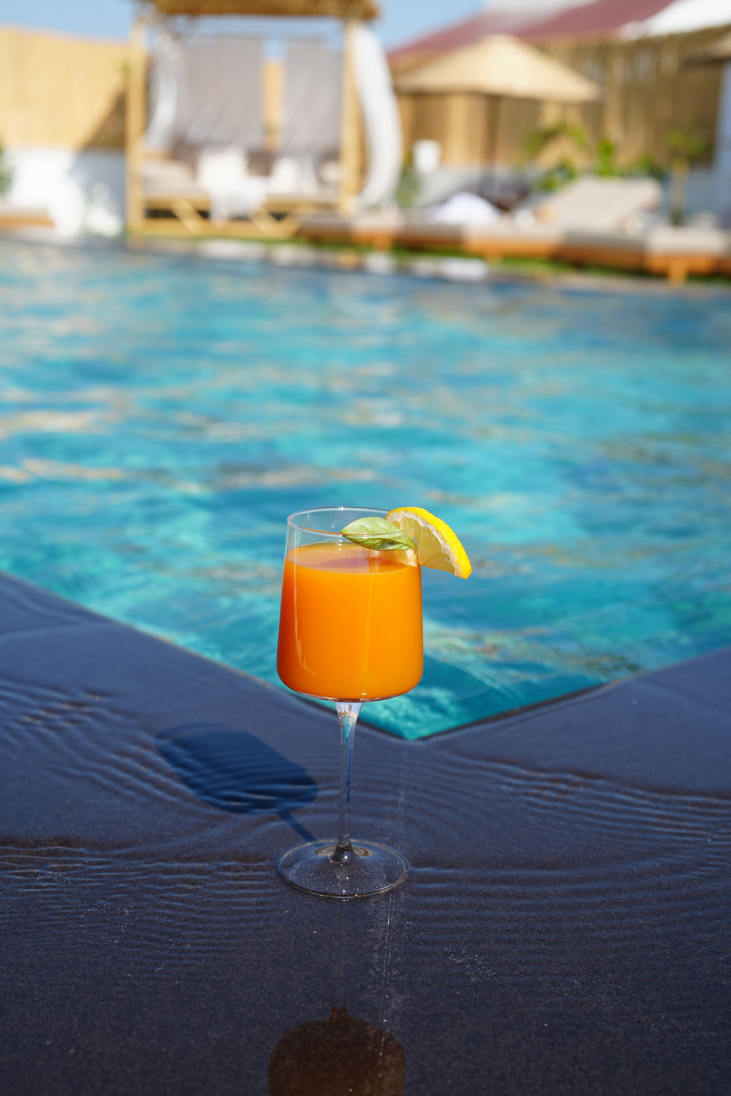 Un verre de boisson à l’orange, garni d’une tranche de citron, posé sur une chaise longue au bord d’une piscine.