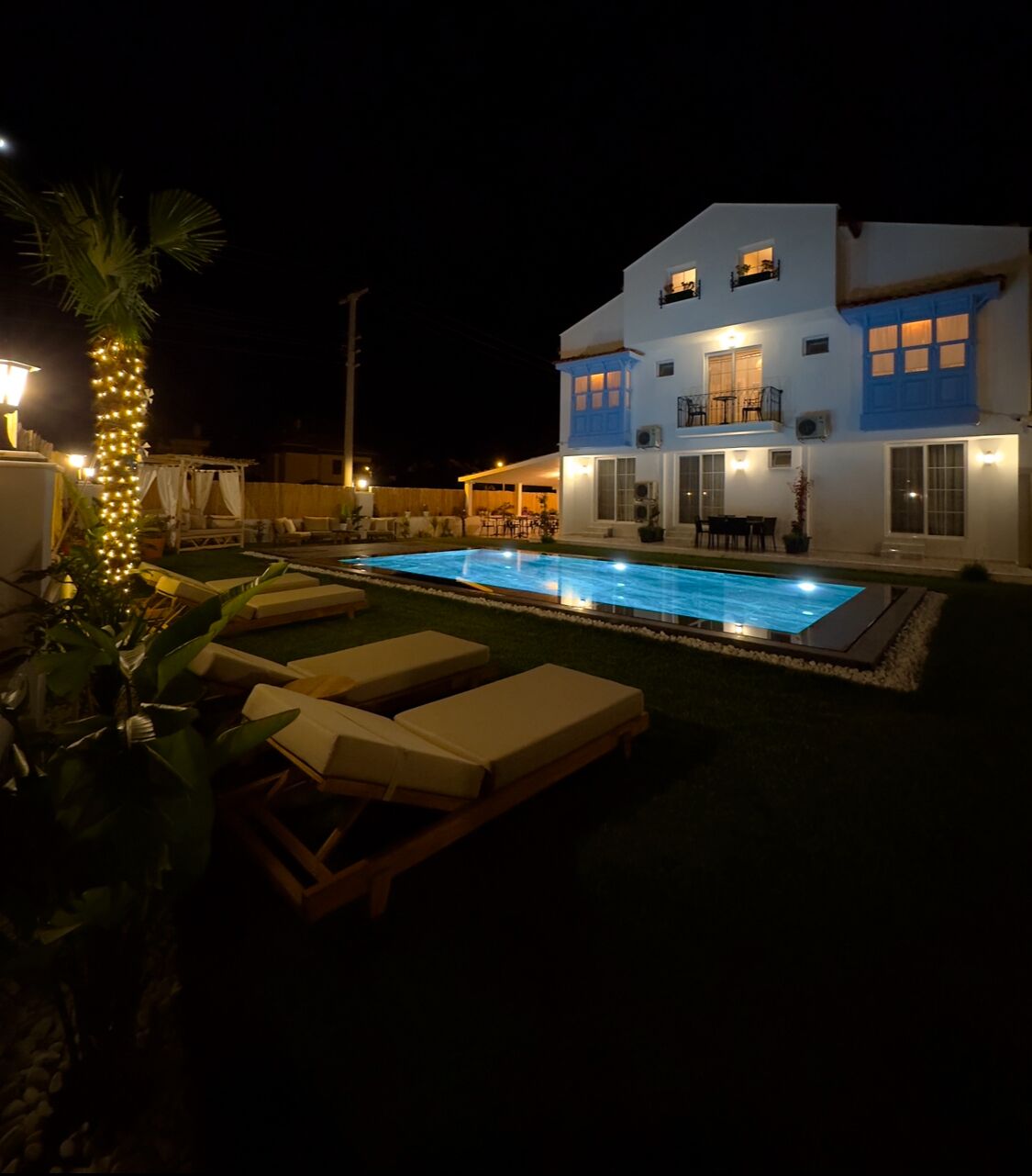 Ein zweistöckiges Haus mit Swimmingpool, Liegestühlen und einem gut beleuchteten Patio.