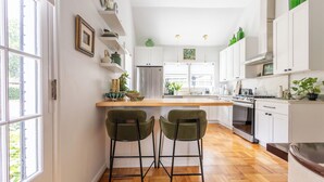 Una cocina moderna con armarios blancos, un refrigerador de acero inoxidable y piso de madera. Hay dos taburetes verdes y un ventanal que proporciona luz natural.