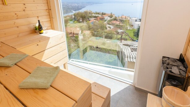 Ein Zimmer aus Holz mit einem großen Fenster mit Blick auf eine Stadt und Wasser. Holzbanken und ein Sauna-Bereich mit Steinen sind vorhanden.