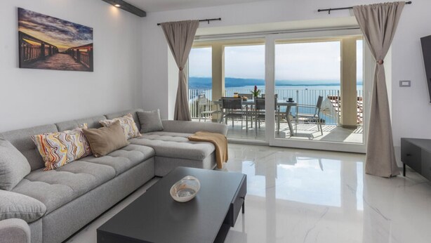 Een moderne woonkamer met een grijze modulaire bank, een zwarte salontafel en een groot raam met uitzicht op zee.