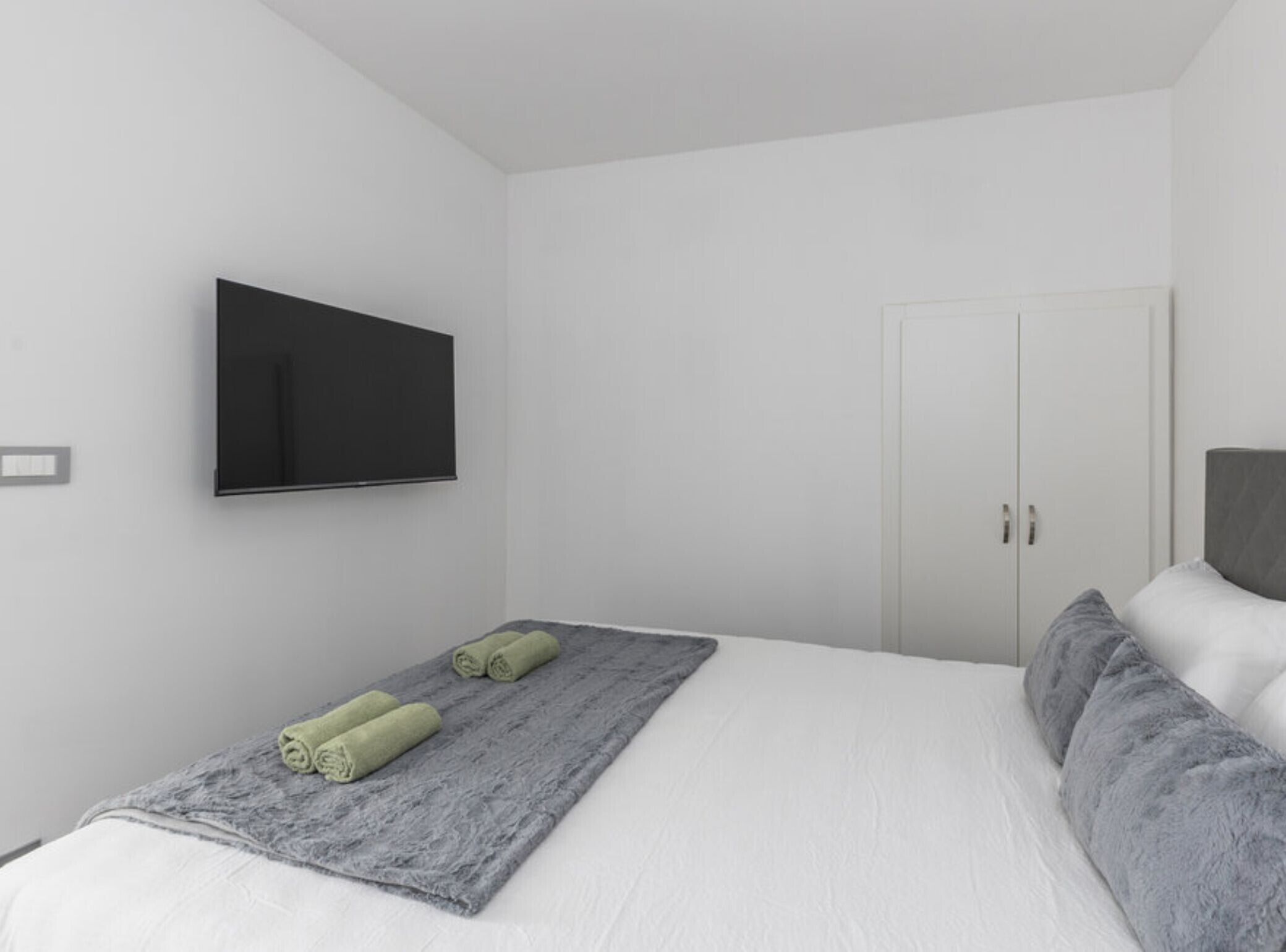 A minimalist bedroom with a bed, a grey blanket, and two rolled towels.