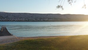 Vista al agua con un muelle que se adentra en aguas tranquilas, un primer plano con césped y un pueblo al fondo.