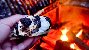 Une main tenant un dessert à la croûte caramélisée, vraisemblablement une crème brûlée, au-dessus d’un feu de camp.