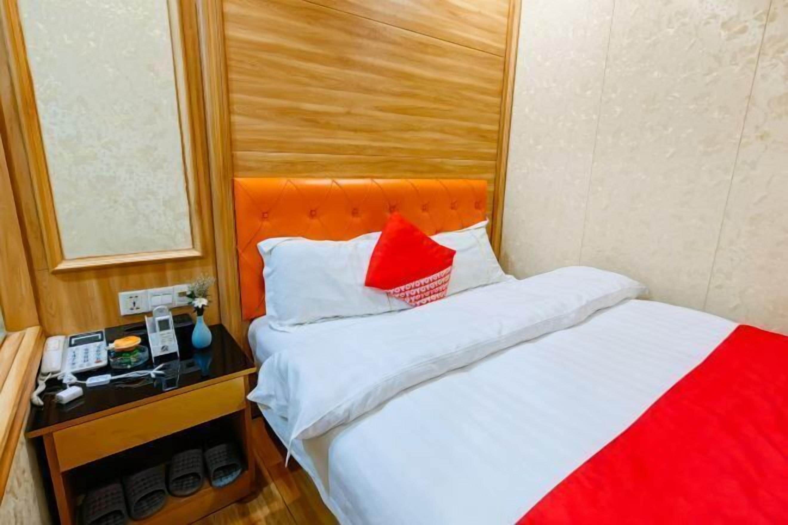 A hotel room with a wooden headboard, a bed with white bedding and a red accent pillow, a bedside table with a phone, a vase, and slippers.