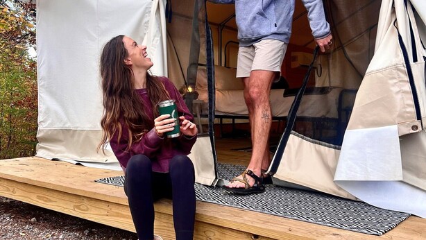 A tent with a wooden platform and a person standing inside, another person sitting outside holding a mug. - Luxury Spacious Glamping, Lake View, Grill, Pool, Pet Friendly, Swim (Delta)