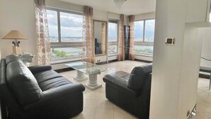 Un salon moderne avec deux canapés en cuir noir, une table basse en verre et un lustre. De grandes fenêtres offrent une vue sur la ville.