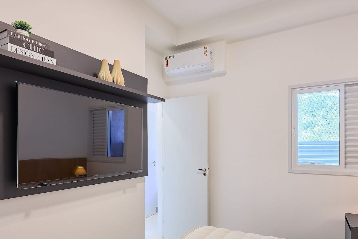 A bedroom with a large mirror, a bedside table with a lamp, a window with blinds, and an air conditioning unit.