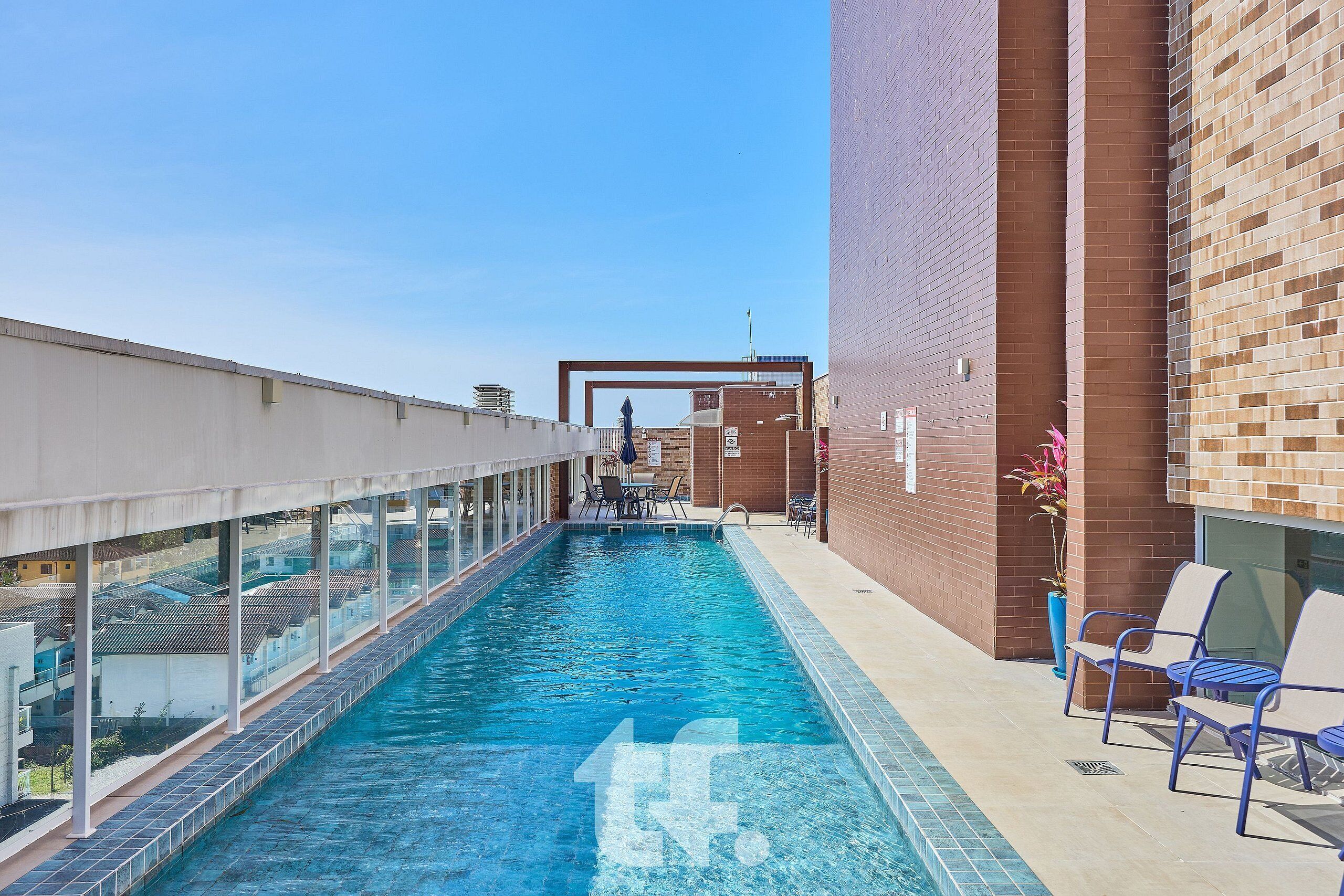 A swimming pool with clear blue water, surrounded by a glass railing and a brick wall. There are lounge chairs and a table with chairs nearby.