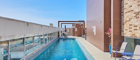 A swimming pool with clear blue water, surrounded by a glass railing and a brick wall. There are lounge chairs and a table with chairs nearby.
