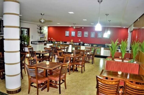 A restaurant with wooden tables and chairs, a red wall with framed pictures, and ceiling fans. - Hotel Arlen 4 Lago Hotel (Lambari)