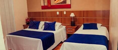 A hotel room with two beds, each with a blue and white bedspread, a wooden headboard, a wall-mounted air conditioner, and a small table with a lamp.