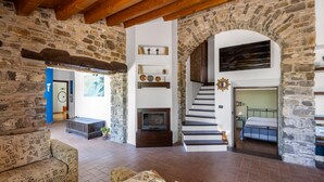 Amplia sala de estar con paredes de piedra, una chimenea, una escalera y una cama en la habitación contigua.