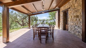 Un patio al aire libre con muro de piedra y vigas de madera, una mesa con sillas y vistas a árboles y agua.