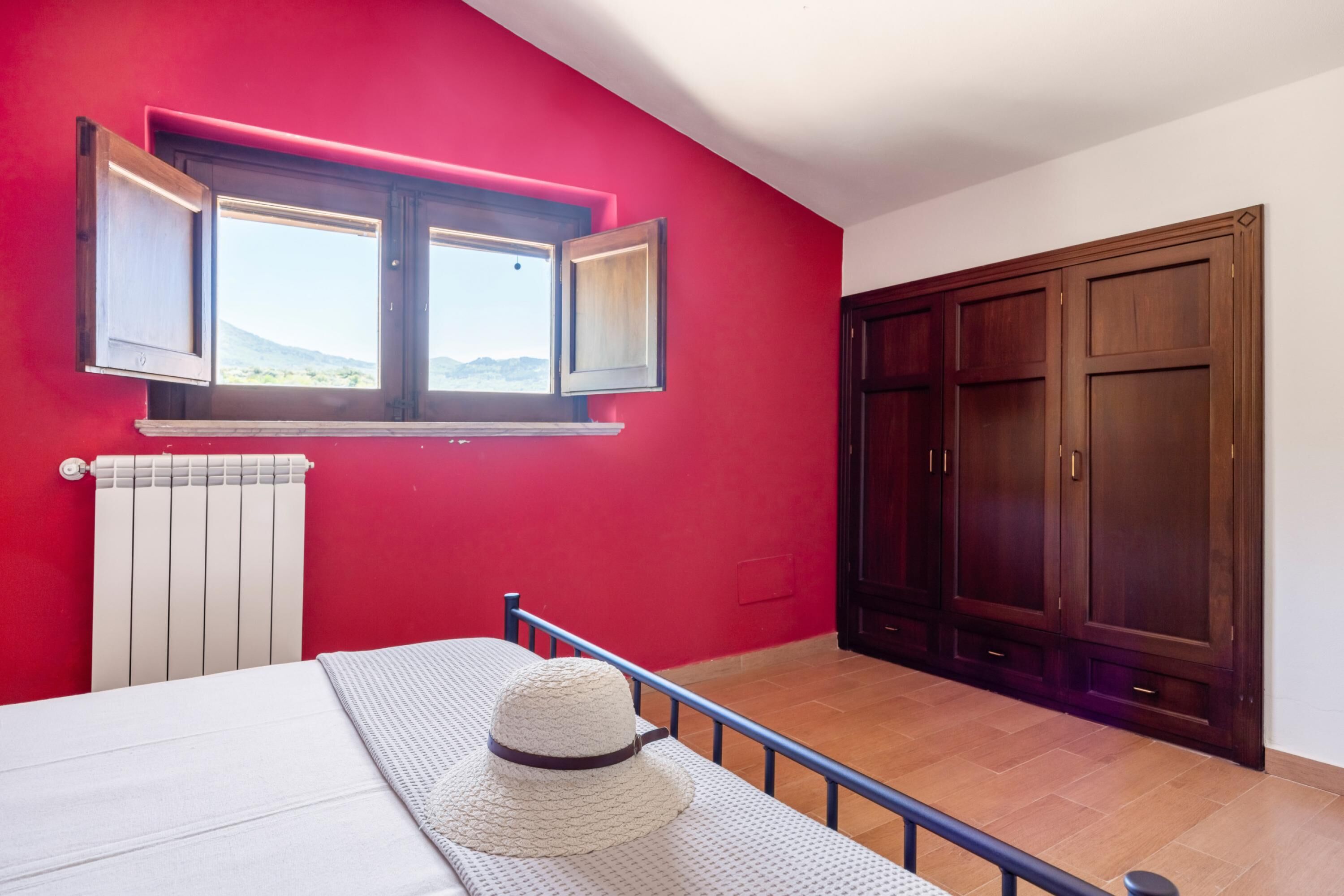 A bedroom with a red wall, a window with open shutters, a radiator, a bed, a hat on the bed, and a wardrobe.
