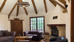 A cozy living room with wooden beams, a fireplace, a sofa, a coffee table, and a window with a view of greenery. - New gîte in vimenet (Aveyron) medieval village between aubrac and lévézou  (Vimenet)