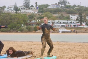 Une scène de plage où l’on voit des personnes en combinaisons de plongée, certaines debout sur des planches de surf, et d’autres allongées sur le sable.