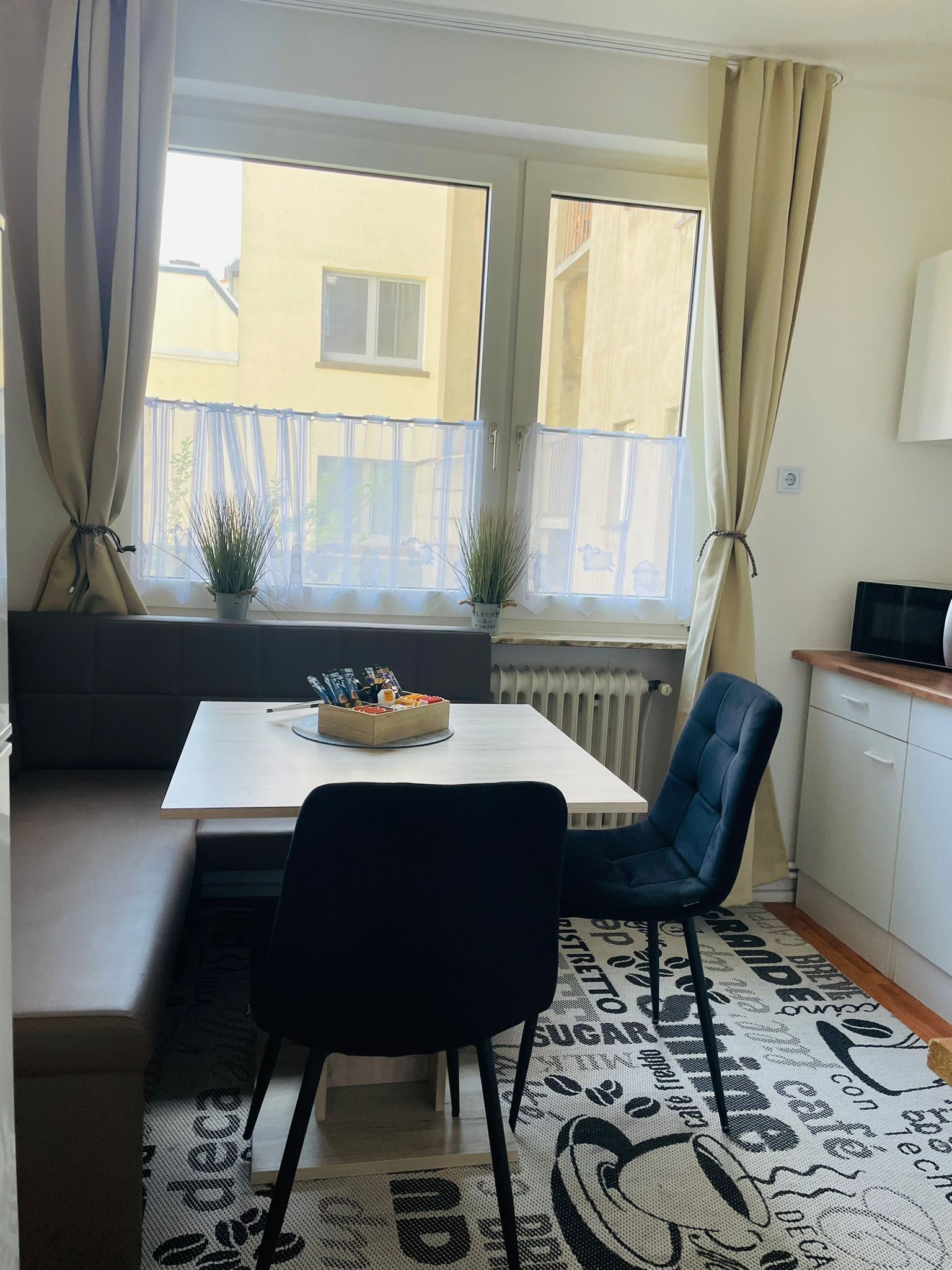 Une cuisine moderne avec un plan de travail blanc, une chaise noire et une table à manger avec un banc. Deux plantes en pot sont posées sur le rebord de la fenêtre et un radiateur se trouve sous les fenêtres.