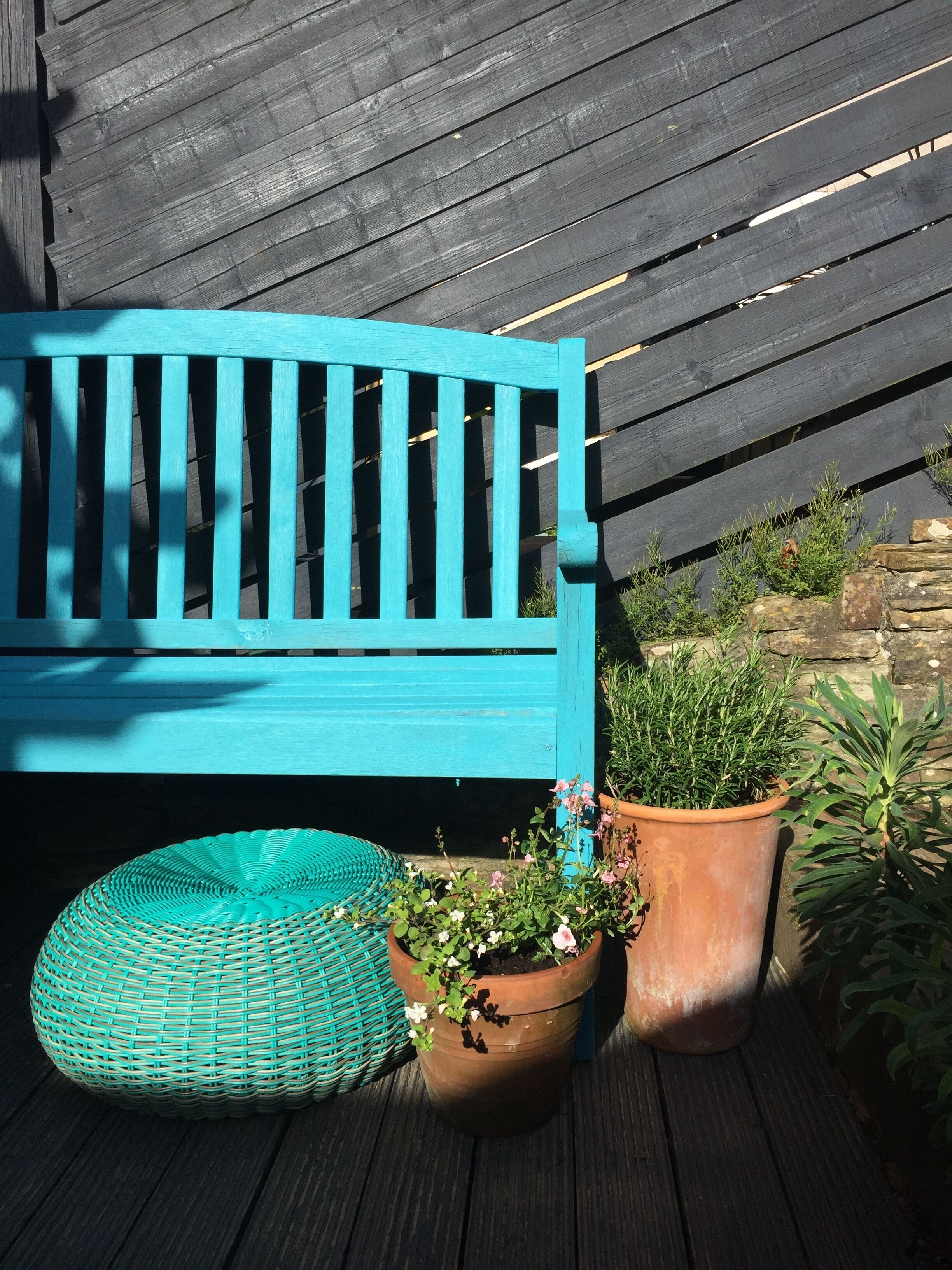 A blue bench, two potted plants, and a woven stool on a wooden deck.