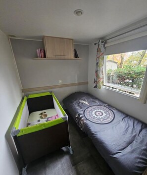 A small room with a bunk bed, a desk with a chair, a window with a view of trees, and a shelf with books.