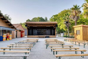 An outdoor amphitheater with a stage, surrounded by wooden benches and a food stall.