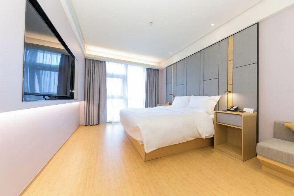 A modern hotel room with a large bed, a bedside table with a lamp, and a view of the outdoors through a window.