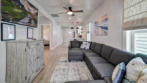 A modern living room with a gray sectional sofa, a wooden cabinet, and a large painting of a mountain landscape. - Nautical Nest - 1 block to the Beach, Newly Renovated (Galveston)