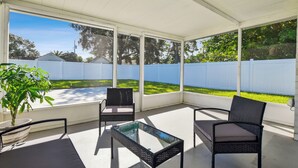 Un patio couvert comprenant deux fauteuils en osier et une table en verre, de grandes fenêtres offrant une vue sur un jardin avec une clôture blanche et de la verdure.
