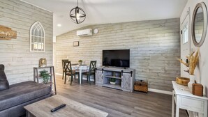Un salon avec un canapé gris, une table basse en bois, un téléviseur fixé à un mur en bois, un coin repas avec une table et des chaises, et une console blanche ornée d’un vase et d’un panier.