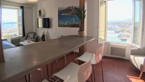 Una cocina moderna con una encimera larga, sillas blancas, un televisor montado en la pared y un ventanal con vistas a un puerto deportivo.