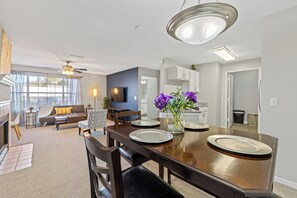 A modern dining area with a wooden table, chairs, and a vase of flowers. There is a ceiling fan and a kitchen in the background.