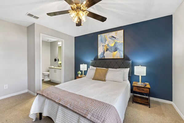 A bedroom with a large bed, a ceiling fan, a nightstand with a lamp, and a painting on the wall.