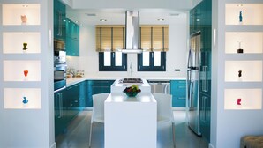 Eine moderne Küche mit türkis-blauen Schränken, weißen Arbeitsplatten und einer zentralen Kochinsel. Es sind Einbaugeräte vorhanden, darunter ein Kühlschrank und ein Backofen, sowie ein Fenster mit Jalousien.