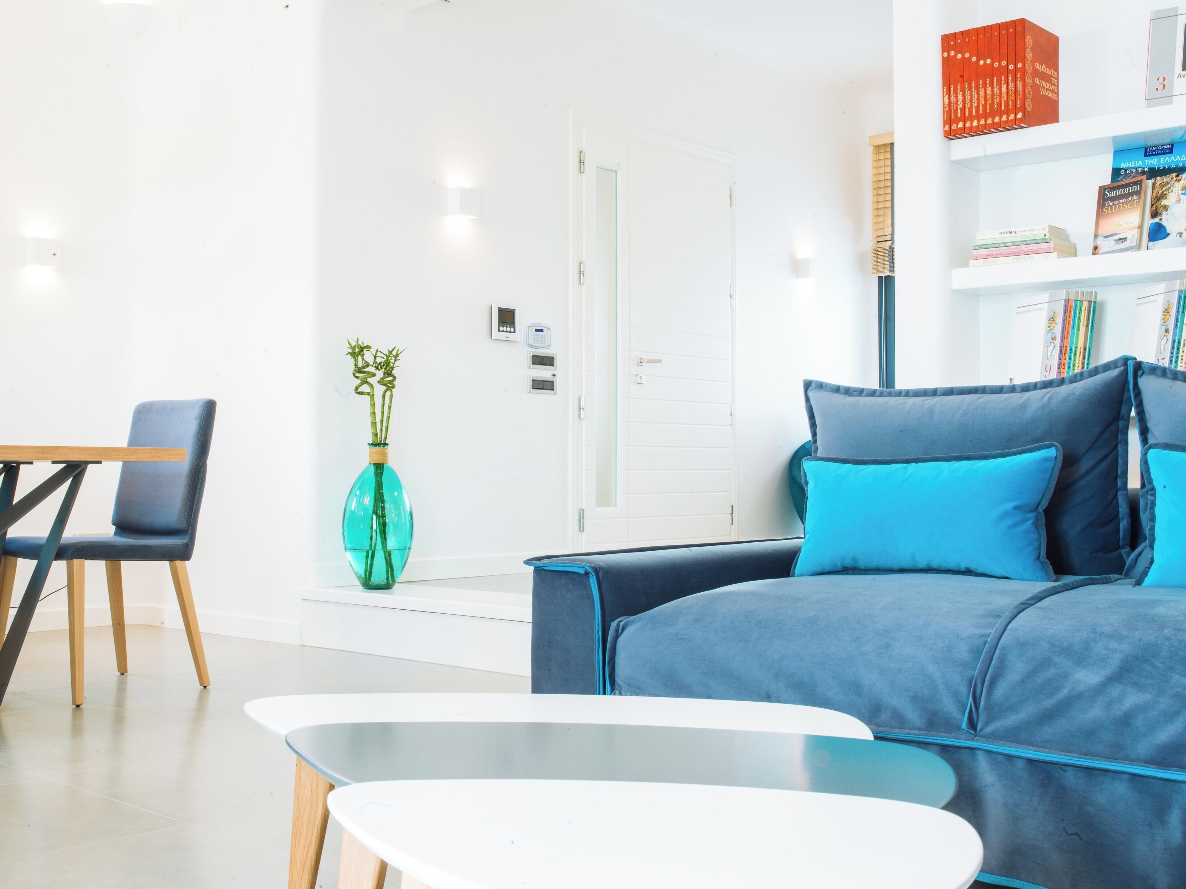 Ein modernes Wohnzimmer mit einem blauen Sofa, einem kleinen Tisch und einem Sessel. An der Wand befindet sich ein Bücherregal mit Büchern und eine Vase mit Grünpflanzen.