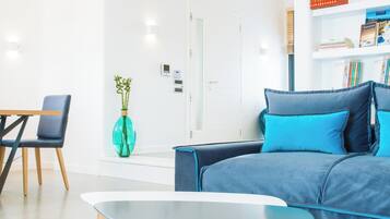 Ein modernes Wohnzimmer mit einem blauen Sofa, einem kleinen Tisch und einem Sessel. An der Wand befindet sich ein Bücherregal mit Büchern und eine Vase mit Grünpflanzen.