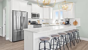 A modern kitchen with white cabinets, stainless steel appliances, a central island with bar stools, and geometric pendant lighting. - Luxury Ocean View Oasis - Private Pool, Hot Tub, Elevator, 100 Steps to Sand! (Ocean Isle Beach)