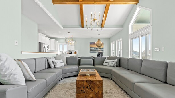 A modern living room with a sectional sofa, a wooden coffee table, and a chandelier. - Luxury Ocean View Oasis - Private Pool, Hot Tub, Elevator, 100 Steps to Sand! (Ocean Isle Beach)