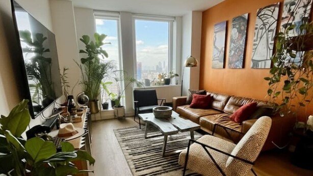 Een moderne woonkamer met een bank, salontafel en stoelen, grote ramen met uitzicht op de stad, en diverse potplanten.