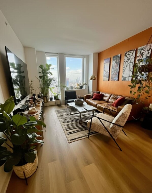 A modern living room with a sofa, coffee table, and chairs, large windows with a city view, and several potted plants.