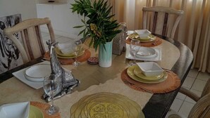 Un comedor con una mesa redonda dispuesta para cuatro personas, rodeada de sillas. Hay una planta en maceta en el centro, y la habitación tiene una ventana con cortinas.