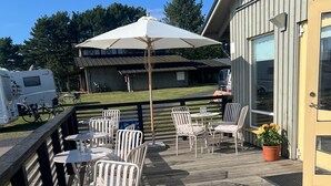 An outdoor deck with white metal chairs and tables, a large umbrella, and a building in the background. - Visit Örestrand (Strandbaden)