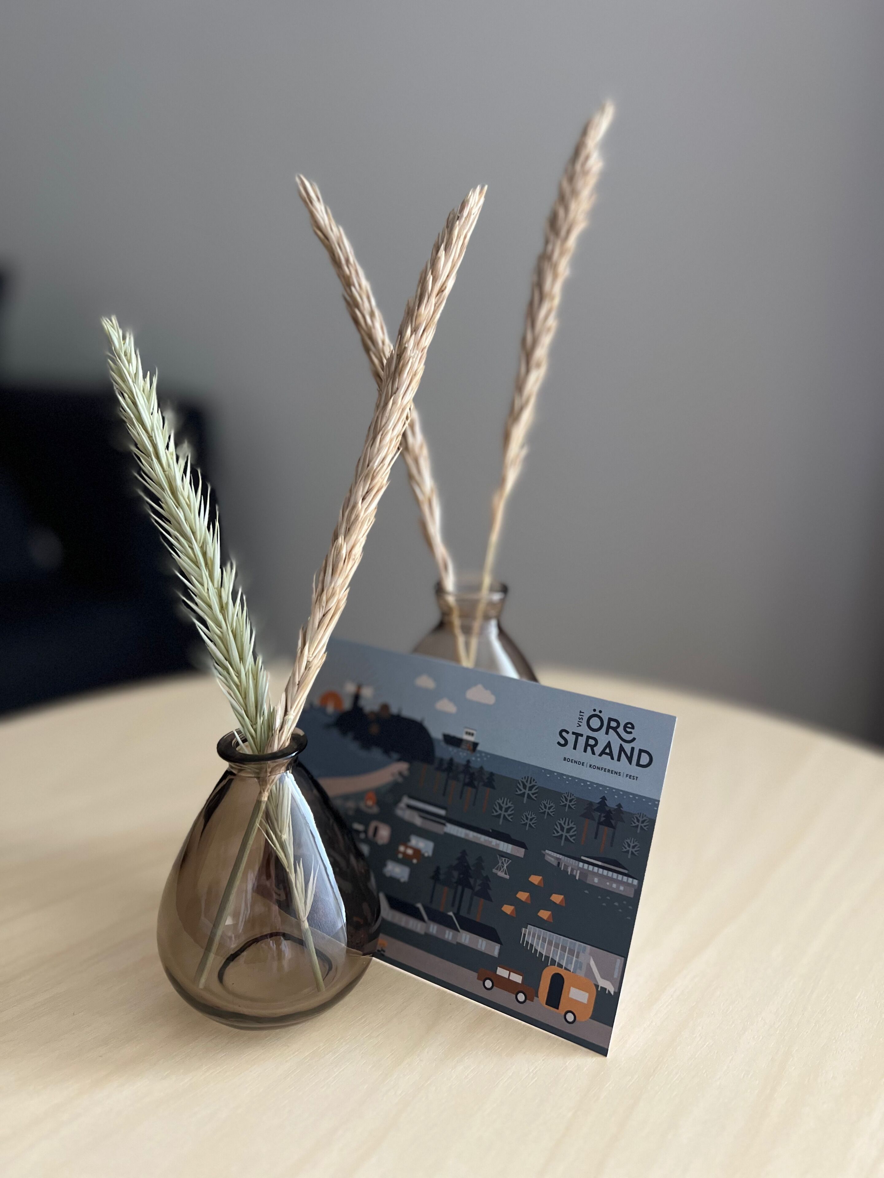 A glass vase with wheat stalks and a brochure titled "Löre Strand" on a wooden surface.