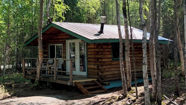 Een houten chalet met een veranda, schoorsteen en een bank, gelegen in een bebost gebied.