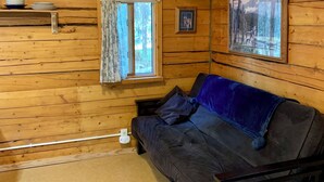 Een gezellig interieur van een chalet met een houten plafond, een bank met een blauwe deken, een klein tafeltje met borden, en lijstjes met foto’s aan de muren.