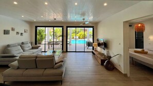 Un vaste espace de vie comprenant un canapé, une table basse et un téléviseur fixé au mur. Une grande fenêtre offre une vue sur une piscine et sur la verdure environnante.